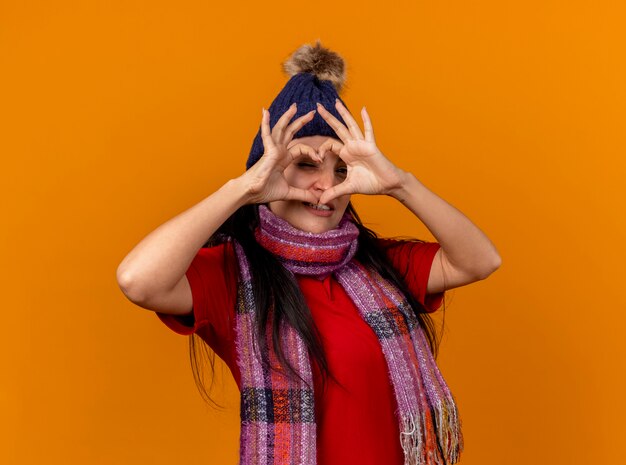 Confident young ill woman wearing winter hat and scarf doing heart sign looking at front through it isolated on orange wall