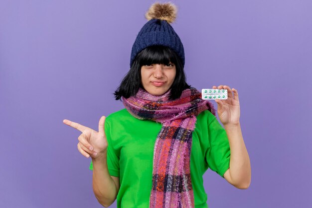 Confident young ill woman wearing winter hat and scarf looking at front holding pack of capsules pointing at side isolated on purple wall with copy space