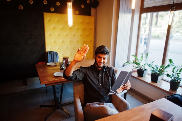 Confident young indian man in black shirt sitting at cafe read menu and calling waiter