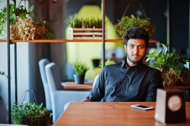 Уверенный молодой индийский мужчина в черной рубашке сидит в кафе