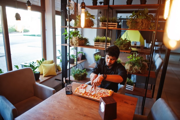 Confident young indian man in black shirt sitting at pizzeria with pizza