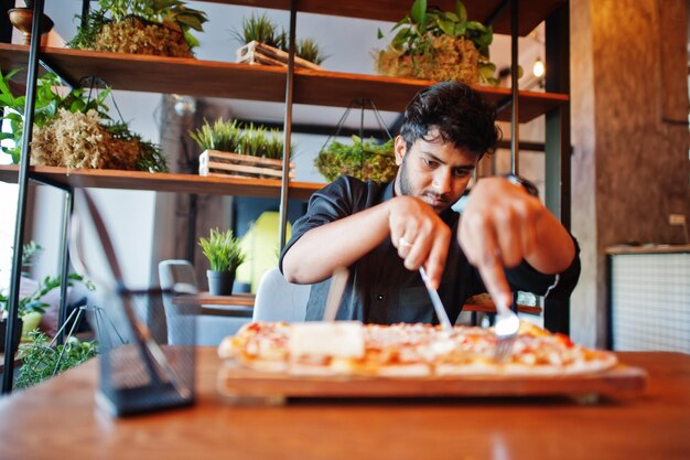 Уверенный молодой индиец в черной рубашке сидит в пиццерии с пиццей