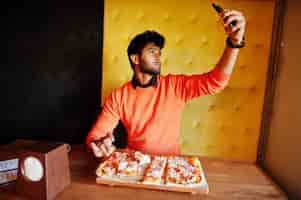 無料写真 ピッツェリアに座っているオレンジ色のセーターを着た自信を持って若いインド人がピザを食べて、彼の携帯電話で写真を撮る