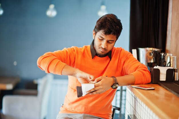 Уверенный в себе молодой индиец в оранжевом свитере сидит за барной стойкой в кафе и рассчитывается с барменом