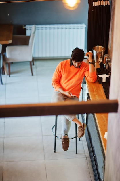 Уверенный в себе молодой индиец в оранжевом свитере сидит за барной стойкой в кафе с мобильным телефоном под рукой