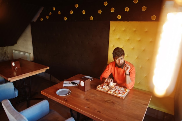 Уверенный молодой индиец в оранжевом свитере сидит в пиццерии и ест пиццу