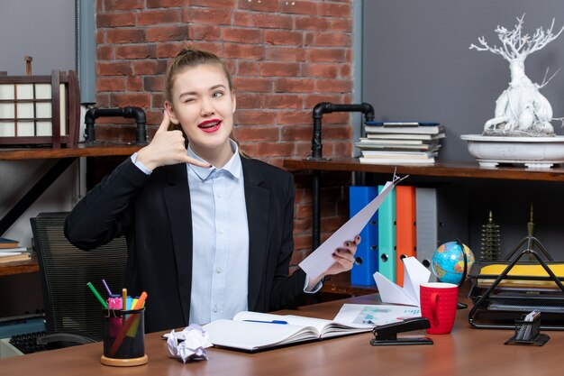 Уверенная в себе молодая леди сидит за столом и держит в руках документ, делая жест "позвони мне" в офисе