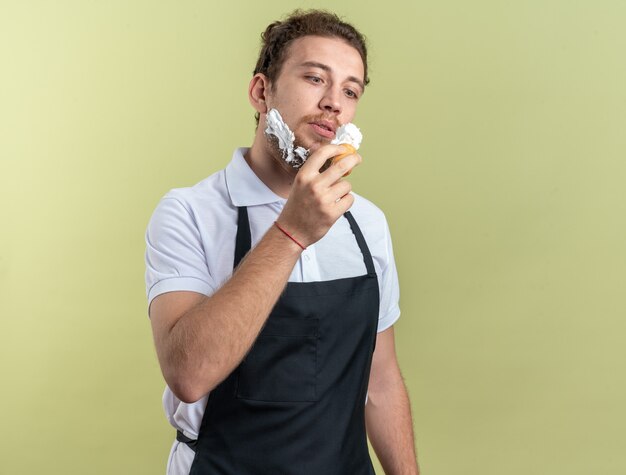 Confident young male barber wearing uniform applied shaving cream with shaving brush isolated on olive green wall