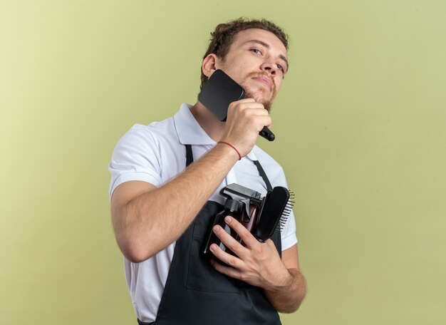 Confident young male barber wearing uniform holding barber tools combing beard isolated on olive green wall