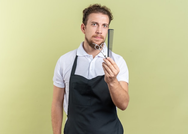 Confident young male barber wearing uniform holding out sciccors with comb isolated on olive green wall