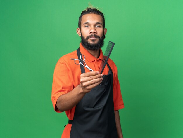 Confident young male barber wearing uniform looking at front stretching out scissors and comb towards front isolated on green wall with copy space