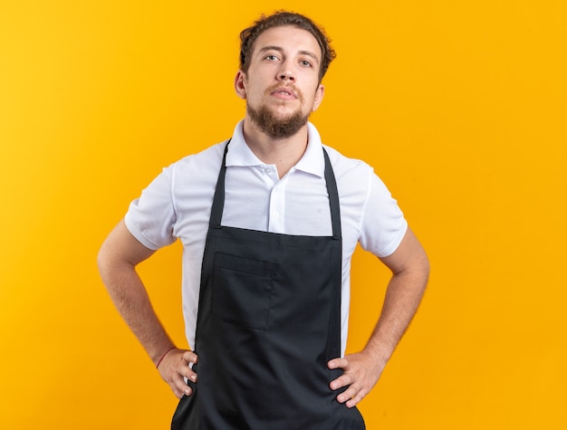 Free photo confident young male barber wearing uniform putting hands on hip isolated on yellow wall