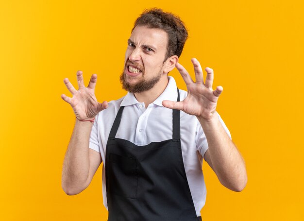 Confident young male barber wearing uniform showing tiger style gesture isolated on yellow wall