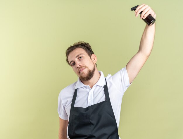 Confident young male barber wearing uniform watering himself with spray bottle isolated on olive green wall