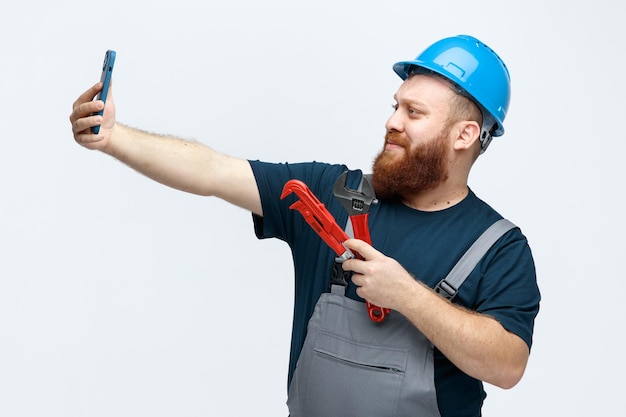 Бесплатное фото Уверенный в себе молодой мужчина-строитель в защитном шлеме и униформе, держащий трубный ключ и гаечный ключ, делающий селфи с мобильным телефоном на белом фоне