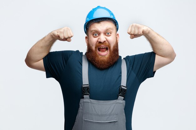 Confident young male construction worker wearing safety helmet and uniform looking at camera showing strong gesture isolated on white background