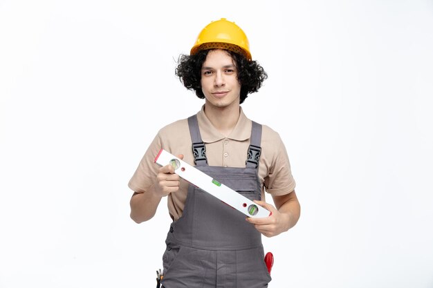 흰색 배경에 격리된 주머니에 건설 도구가 들어 있는 카메라를 바라보고 있는 제복과 안전 헬멧을 쓴 자신감 있는 젊은 남성 건설 노동자