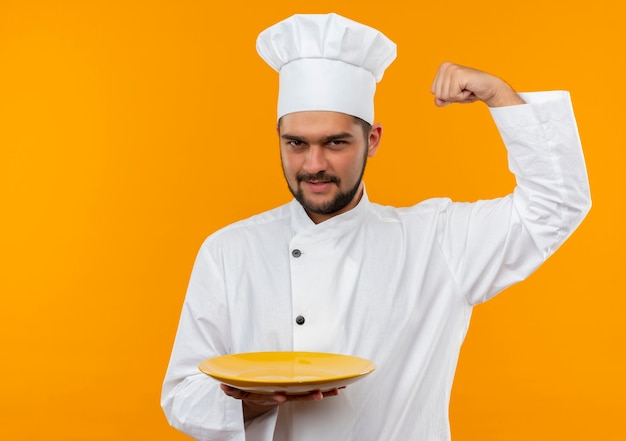 Confident young male cook in chef uniform holding empty plate gesturing strong isolated on orange wall