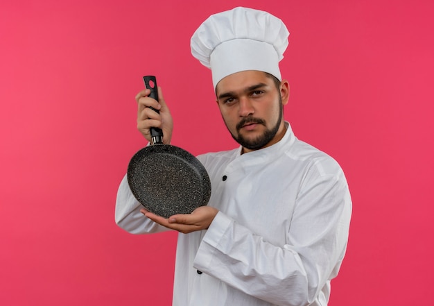 Confident young male cook in chef uniform holding frying pan  isolated on pink wall with copy space