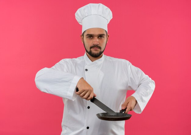 Confident young male cook in chef uniform holding knife and frying pan  isolated on pink wall