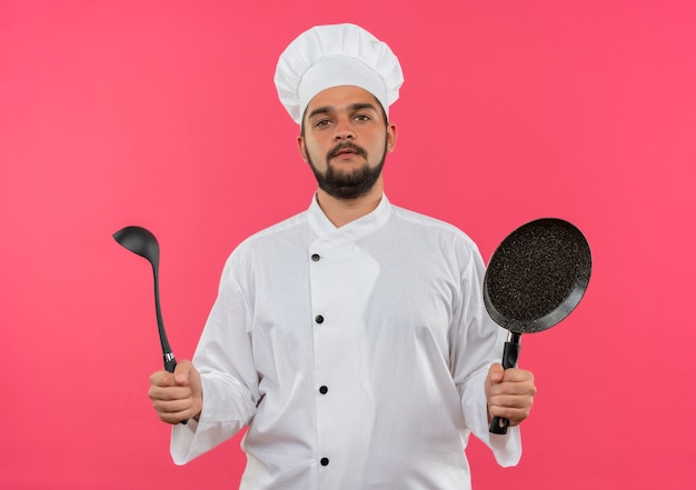 Confident young male cook in chef uniform holding ladle and frying pan  isolated on pink wall