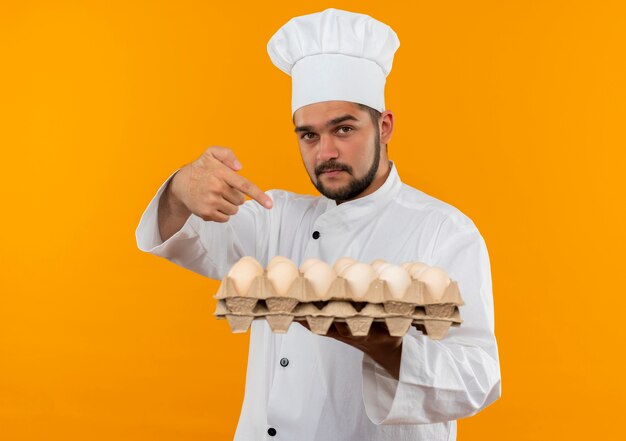 Уверенный молодой мужчина-повар в униформе шеф-повара держит и указывает на коробку яиц, изолированную на оранжевой стене