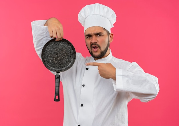 Уверенный молодой мужчина-повар в униформе шеф-повара держит и указывает на сковороду, изолированную на розовой стене