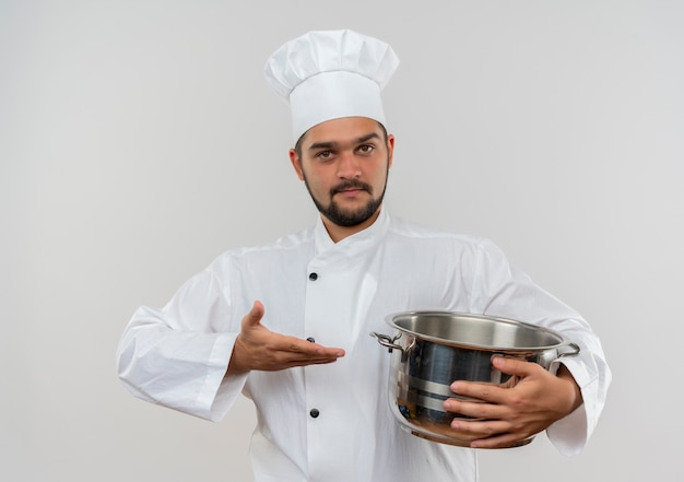 요리사 유니폼 들고 흰색 벽에 고립 된 냄비에 손으로 가리키는 자신감 젊은 남성 요리사