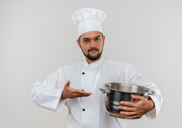 Confident young male cook in chef uniform holding and pointing with hand at pot isolated on white wall