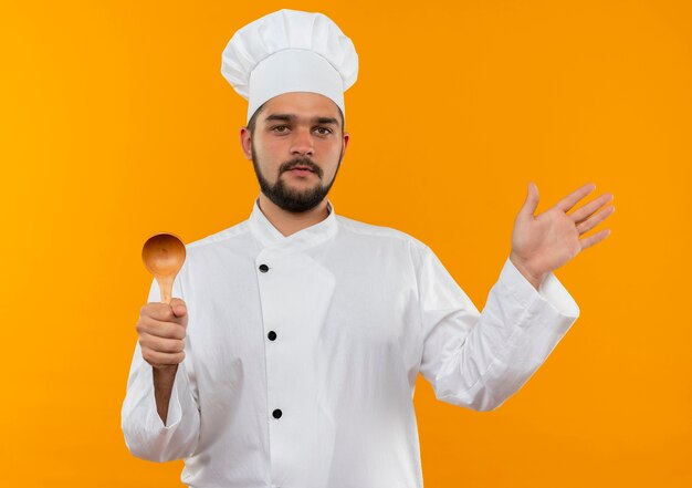 Fiducioso giovane cuoco maschio in uniforme da chef tenendo il cucchiaio e mostrando la mano vuota isolata sul muro arancione