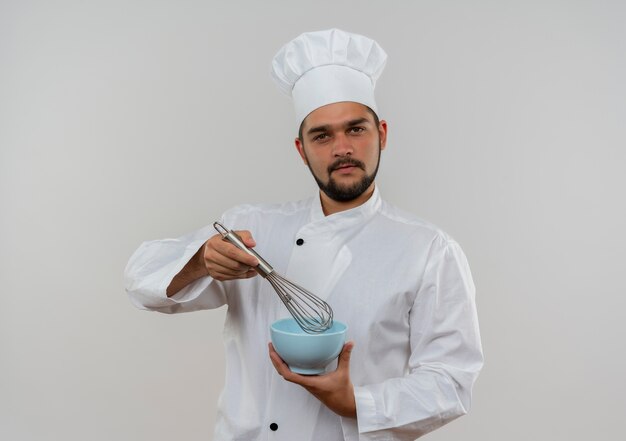 Confident young male cook in chef uniform holding whisk and bowl looking  isolated on white wall with copy space