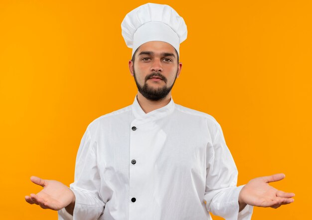 오렌지 벽에 고립 된 빈 손을 보여주는 요리사 유니폼에 자신감이 젊은 남성 요리사