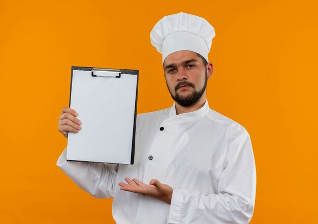 Confident young male cook in chef uniform showing and pointing with hand at clipboard isolated on orange wall