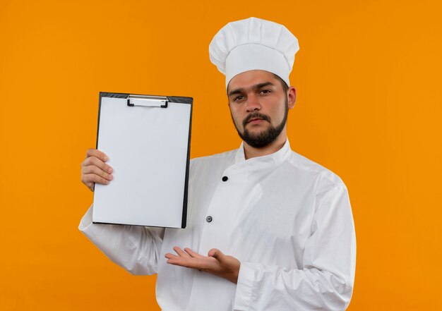 オレンジ色の壁に分離されたクリップボードを示し、手で指しているシェフの制服を着た自信のある若い男性料理人