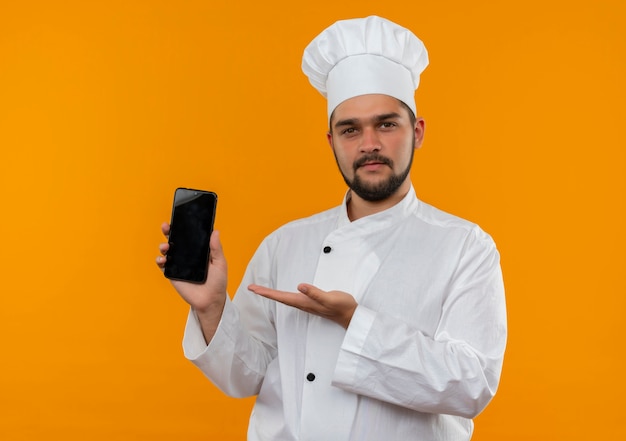 Confident young male cook in chef uniform showing and pointing with hand at mobile phone isolated on orange wall