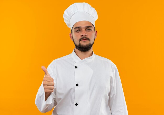 Уверенный молодой мужчина-повар в униформе шеф-повара показывает большой палец вверх изолированным на оранжевой стене