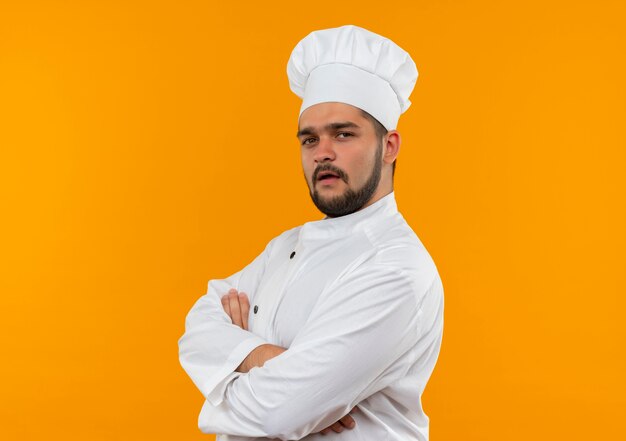 Confident young male cook in chef uniform standing in profile view with closed posture isolated on orange wall with copy space