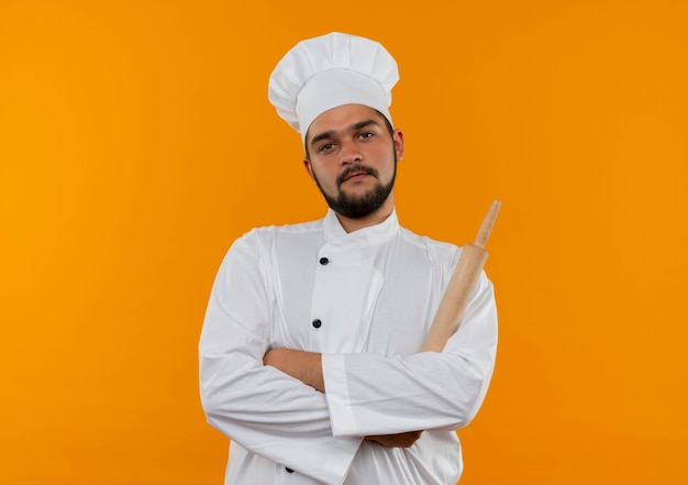 Уверенный молодой мужчина-повар в униформе шеф-повара стоит в закрытой позе, держа скалку на оранжевой стене с копией пространства