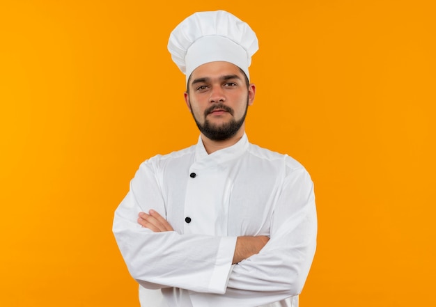 コピースペースのあるオレンジ色の壁に閉じた姿勢で立っているシェフの制服を着た自信のある若い男性料理人