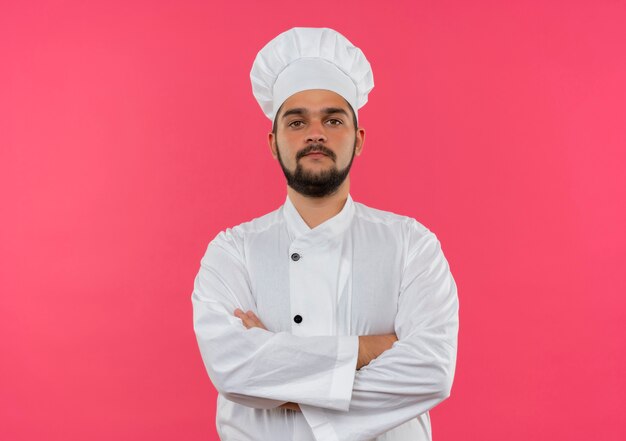 Confident young male cook in chef uniform standing with closed posture  isolated on pink wall with copy space