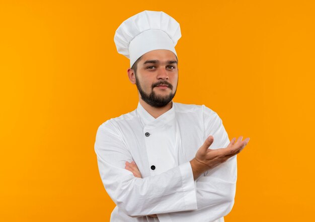 Confident young male cook in chef uniform standing with closed posture and showing empty hand isolated on orange wall with copy space
