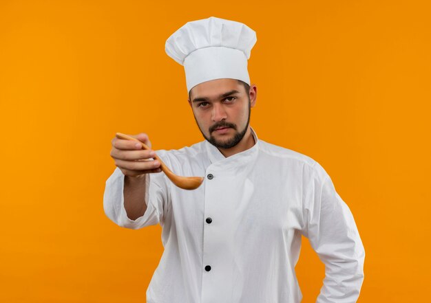 Уверенный молодой мужчина-повар в униформе шеф-повара протягивает ложку, изолированную на оранжевой стене