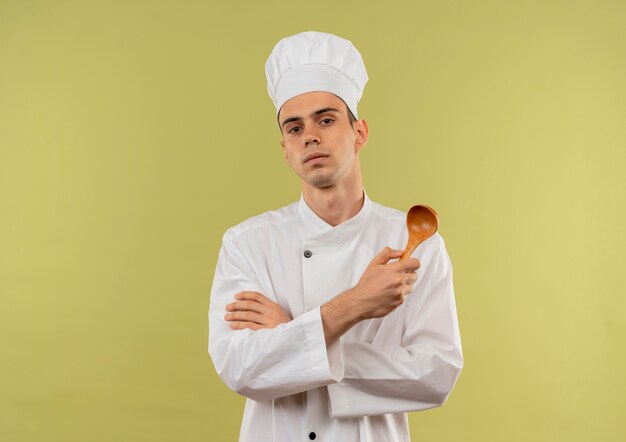 Уверенный молодой мужчина-повар в униформе шеф-повара, скрестив руки, держа ложку на изолированной зеленой стене с копией пространства