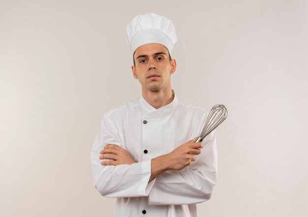 Confident young male cook wearing chef uniform crossing hands holding whisk on isolated white wall with copy space