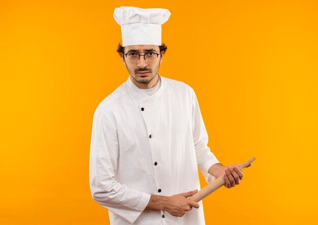 Уверенный молодой мужчина-повар в униформе шеф-повара и в очках держит скалку