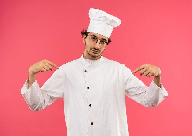 Уверенный в себе молодой повар в униформе шеф-повара и в очках указывает на себя