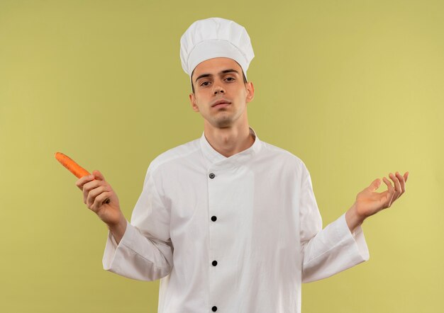 Confident young male cook wearing chef uniform holding carrot and spreads hands on isolated green wall