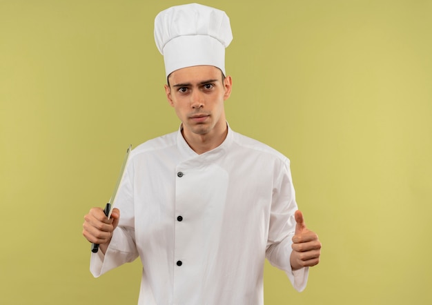 Fiducioso giovane cuoco maschio che indossa l'uniforme del cuoco unico che tiene la mannaia il pollice in su sulla parete verde isolata con lo spazio della copia