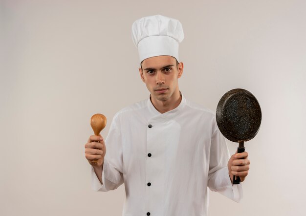 Уверенный молодой мужчина-повар в униформе шеф-повара держит сковороду и спу на изолированной белой стене