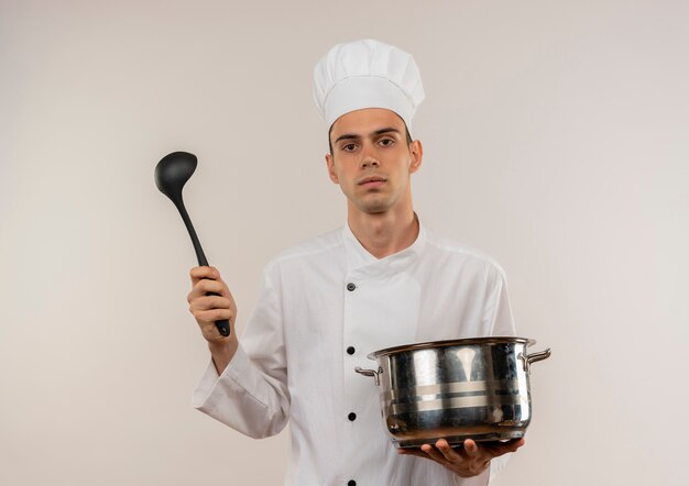 Confident young male cook wearing chef uniform holding saucepan and ladle on isolated white wall